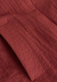 Gros plan sur un tissu en velours côtelé couleur rouille présentant une poche passepoilée rectangulaire avec des coutures apparentes et des nervures texturées.