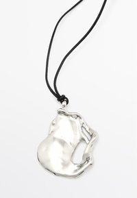 Pendente in argento dalla forma astratta, con una superficie liscia e riflettente. Attaccato a un cordino nero. Il design presenta curve organiche e uno stile minimalista.