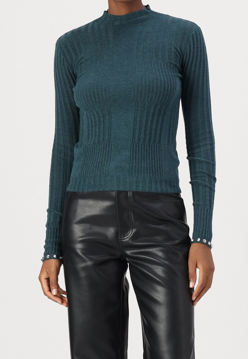 Haut en maille côtelée à manches longues couleur sarcelle avec un col montant, présentant des rayures texturées et des boutons aux poignets, assorti à un pantalon en cuir noir.