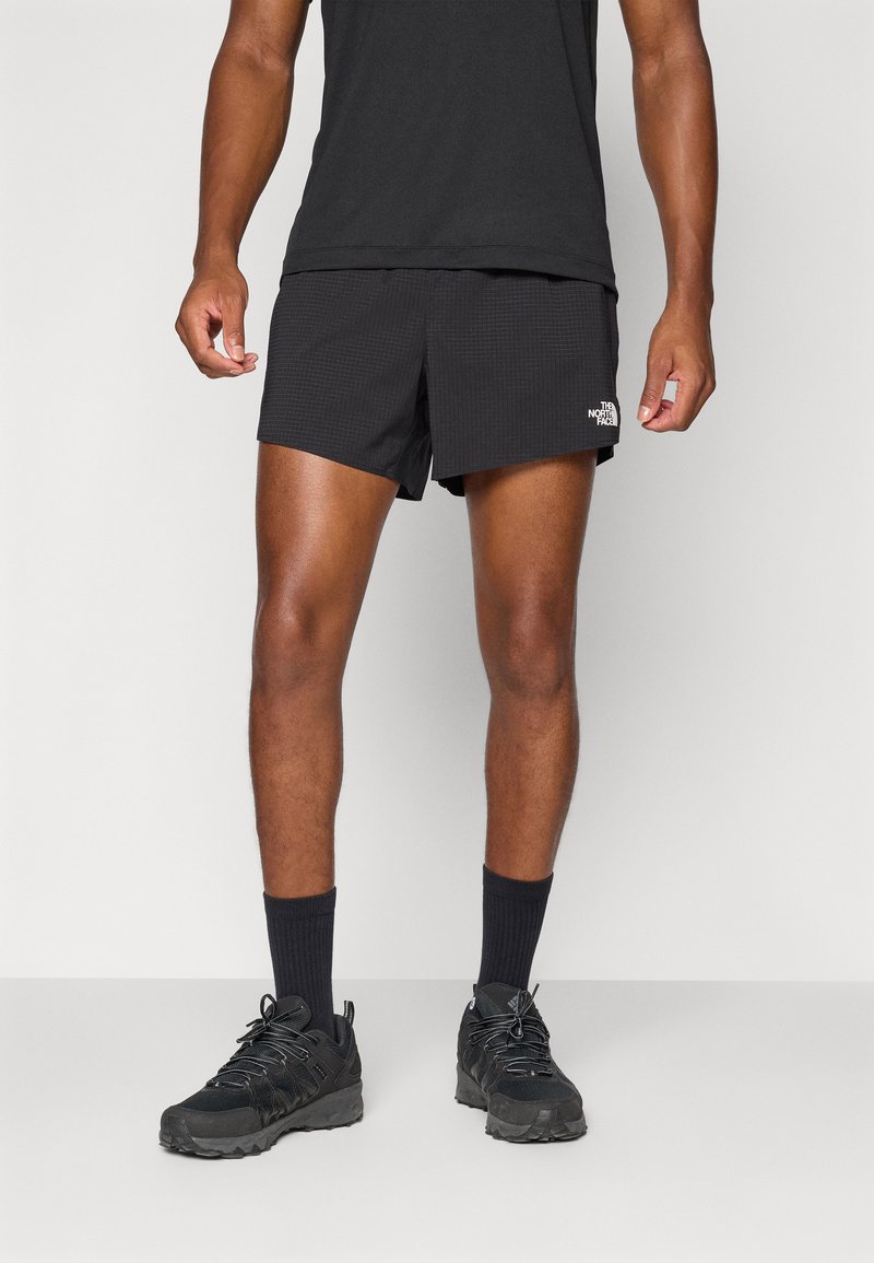 Schwarze Sportshorts mit strukturiertem Muster, ausgestattet mit einem weißen Logo am unteren Bein, kombiniert mit schwarzen Socken und Trailrunning-Schuhen.