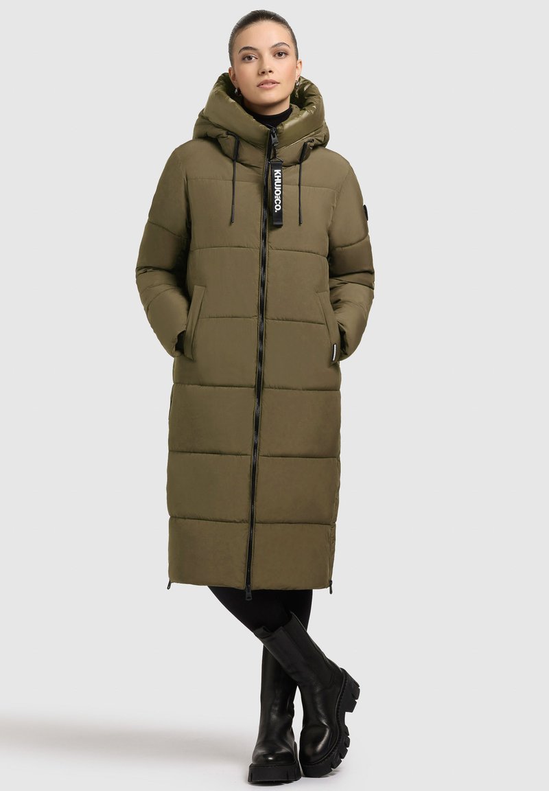 Khaki dlouhý zateplený kabát s kapucí, zapínání na zip, boční kapsy a štítek s logem. Noseno přes černé legíny a boty.