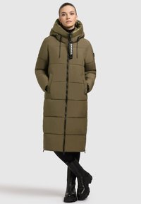 Olijfgroene gewatteerde lange jas met een capuchon, ritssluiting, zijzakken en een merketiket. Draagt over zwarte leggings en laarzen.