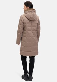 Beige pufferjakke med hætte, quiltet tekstur og længere længde, parret med sorte bukser og kraftige sorte støvler.