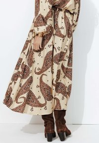 Robe beige à motif paisley avec des manches longues et une taille cintrée, associée à des bottes en suède marron jusqu'aux genoux avec un talon épais.