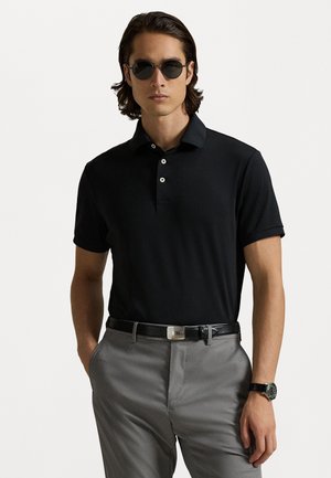 Svart kortermet polo-skjorte med tre knapper, kombinert med grå bukser og et svart belte. Tilbehøret inkluderer runde svarte solbriller og et armbåndsur.