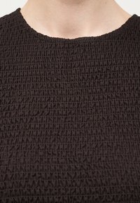 Κοντινό πλάνο στον λαιμό και το πάνω μέρος του στήθους ενός ατόμου που φορά κορυφή από υφή σκούρου καφέ ύφασμα με ραφές σμόκιν και στρογγυλή λαιμόκοψη.