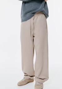 Pantalons beige à jambe large avec une taille élastique et un détail de cordon. Texture du tissu lisse associée à des baskets de teintes neutres.
