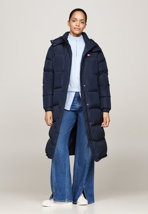Kvinde iført lang navy dunjakke over lyseblå top og brede blå jeans med hvide sneakers, stående mod en ensfarvet baggrund.