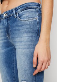 Jeans de denim azul claro con un diseño desgastado, que presentan un ligero deterioro y costuras en contraste. Vista en primer plano de la cintura y la mano.
