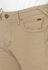 Jeans de mezclilla beige con un corte ajustado, diseño de cinco bolsillos, cierre de botón metálico y una sutil etiqueta de marca en el bolsillo frontal.