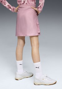 Persona vestida con una falda rosa con un bolsillo lateral, calcetines blancos de Puma y zapatillas blancas de Puma, de pie frente a un fondo de degradado gris.