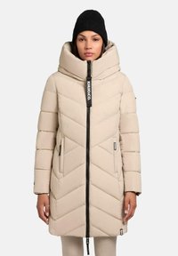 Beige pufferjack met een hoge kraag, een chevronpatroon en een zwarte rits. Inclusief zijzakken en een zwart merklabel. Draagt een zwarte gebreide muts.
