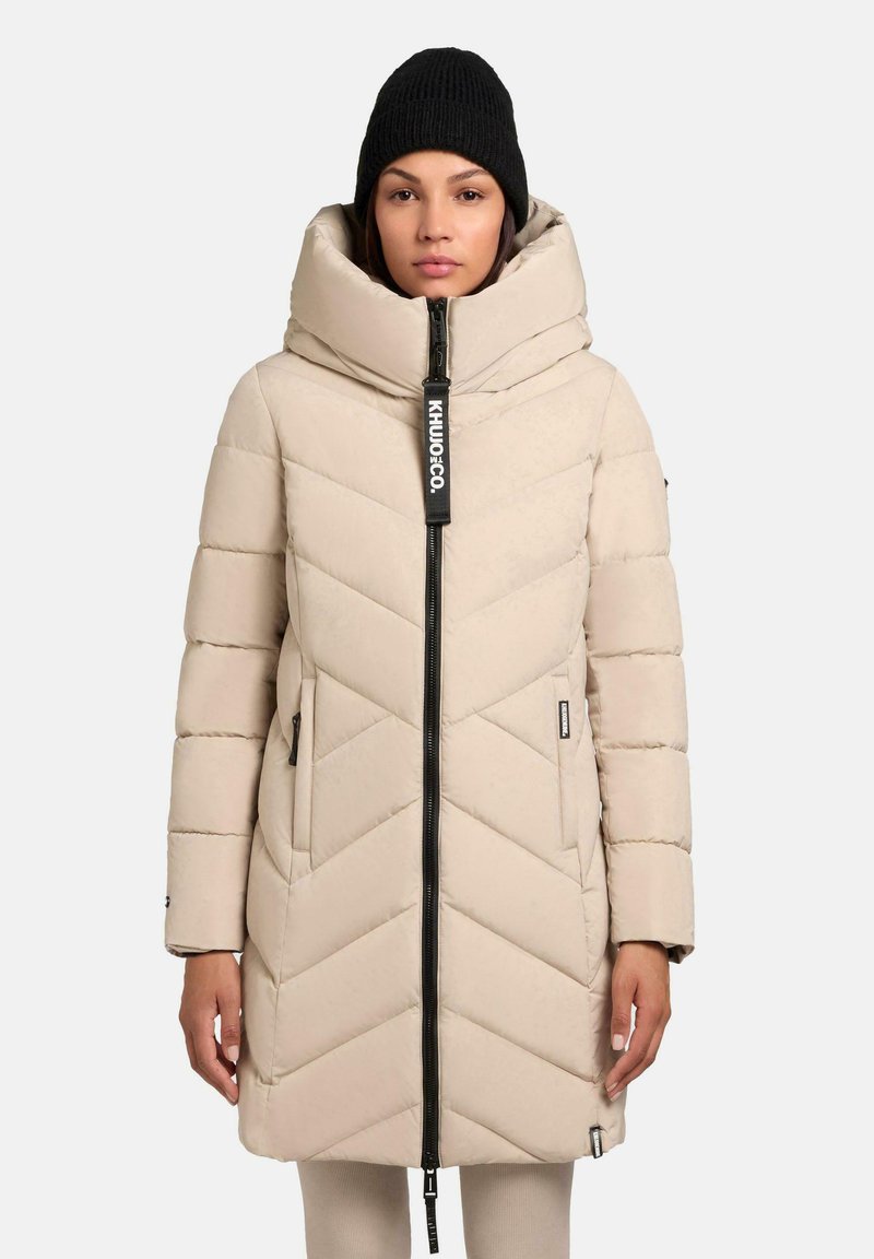 Beige pufferjack met een hoge kraag, een chevronpatroon en een zwarte rits. Inclusief zijzakken en een zwart merklabel. Draagt een zwarte gebreide muts.