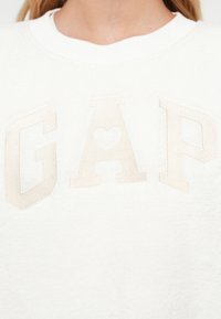 Felpa in pile bianca con la scritta ricamata "GAP" in beige, con una forma a cuore al centro della lettera "A".