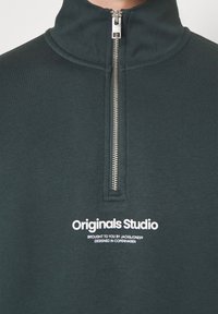 Tmavě zelený svetr s čtvrtzipem, stříbrným zipem a bílým logem "Originals Studio" a menším textem pod ním na hrudi.