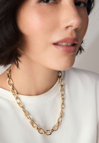 Gold chain necklace with oval links, worn over a white shirt. Smooth, polished finish with a shiny texture. Neatly clasped at the back.