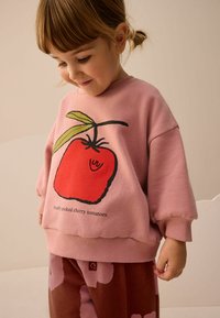 Ružová mikina s veľkou grafikou červenej rajčiny so zeleným lístkom, pod ňou text "Čerstvo zozbierané čerešňové rajčiny," vo voľnom strihu.