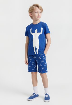 Dreng stående med hænder i lommerne iført blå t-shirt med hvidt figurgrafik, blåmønstrede shorts, hvide sokker og blå sneakers.