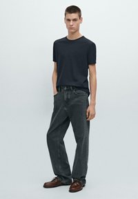 T-shirt nero a maniche corte abbinato a jeans grigi scuri a vita alta e modello ampio. Il modello è in piedi con le mani nelle tasche. Scarpe marroni ai piedi.