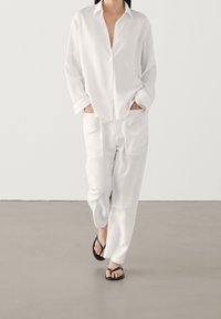 Chemise blanche boutonnée avec manches longues et col, associée à un pantalon blanc ample. Tongs noires. Design minimaliste et texture douce.
