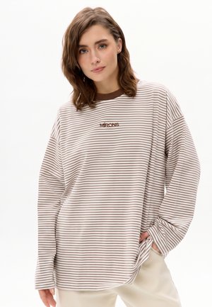 Junge Frau mit schulterlangem braunem Haar, die ein beige-braun gestreiftes Langarmshirt und helle Hosen trägt.
