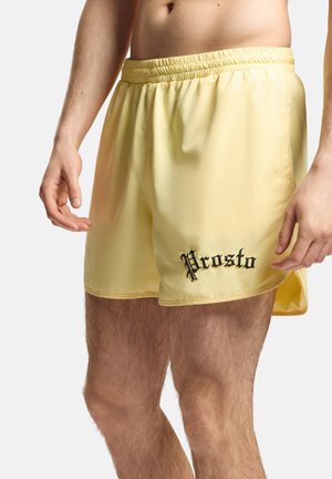 Mężczyzna noszący jasnżółte szorty z elastycznym pasem i czarnym napisem „Prosto” na lewym udzie na jednolitym tle.
