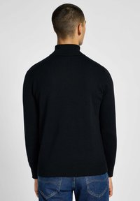 Maglione a collo alto nero con maniche lunghe, colletto e orlo a costine. Indossato con jeans in denim blu. Tessuto di consistenza morbida, design aderente.