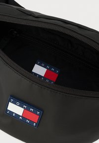 Zwarte stoffen heuptas met ritssluiting; binnenkant voorzien van een blauw, rood en wit merklabel. Gladde textuur, compact ontwerp.