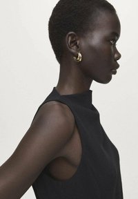 Zwart mouwloos topje met een hoge halslijn. Kort, getextureerd haar en gouden oorringen. Zijaanzicht met nadruk op gelaatstrekken en details.