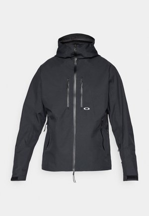 Chaqueta impermeable negra con capucha, cierre frontal, bolsillos con cremallera en el pecho y los laterales, y correas ajustables en las muñecas.