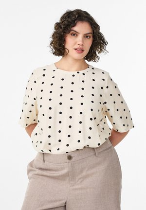 Kobieta o krótkich kręconych włosach, ubrana w białą bluzkę w czarne kropki i beżowe spodnie, stojąca z rękami za plecami.