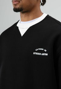Zwart sweatshirt met een v-hals ontwerp, voorzien van een witte ondershirtkraag. Bevat witte tekst: "LE-75005-DE STUDIO ABYSS." Zachte textuur.