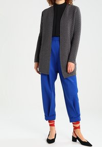 Szary, teksturowany kardigan z otwartym przodem, połączony z niebieskimi spodniami i czerwonymi skarpetkami w paski. Całość dopełniają czarne, spiczaste baleriny.