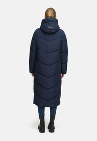 Lange marineblauwe pufferjas met capuchon, met chevron-quilting, een logo bij de nek, en gedragen over denim en zwarte laarzen.