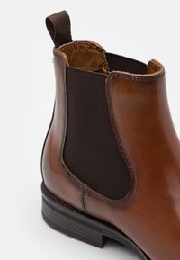 Bottine en cuir marron avec panneaux latéraux élastiques, languette d'aide à l'enfilage et détails noirs texturés. Semelle robuste avec légère empreinte.