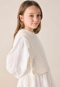 Strickweste in Creme mit strukturiertem Muster, weicher Oberfläche und Rippensaum, getragen über einem weißen Kleid mit Puffärmeln und floraler Stickerei.