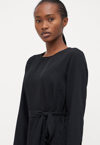 Μαύρο φόρεμα με μακριά μανίκια, κατασκευασμένο από υφή υφάσματος, με στρογγυλή λαιμόκοψη και ζώνη στη μέση που δένει για ρυθμιζόμενη εφαρμογή.