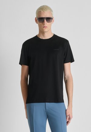 Hombre rubio con camiseta negra, pantalones azules, gafas de sol oscuras de gran tamaño, de pie frente a un fondo liso gris claro.