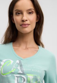 Hellgrünes V-Ausschnitt-T-Shirt aus weichem Stoff mit grafischem Text und Mustern in Weiß, Grau und Grün. Nahaufnahme.