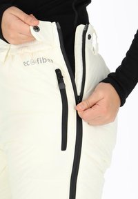 Händer som drar upp en vit jacka med svart dragkedja och tryckknapp, som visar texten "Eco Fiber" på tyget.