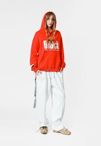 Sudadera roja con capucha que presenta un gráfico de "Black" y un diseño de oso, combinada con pantalones blancos de gran tamaño y zapatos beige, con acentos verdes.