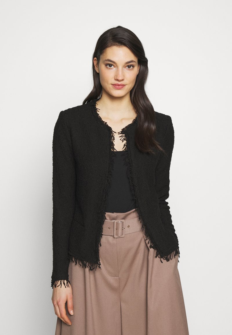 Veste noire texturée avec des bords effilochés, col rond et devant ouvert. Stylisée par-dessus un top noir et un pantalon taille haute beige.