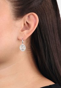 Orecchino a goccia rotondo in argento con un intricato design a foglia, indossato sull'orecchio sinistro di una persona con capelli castano scuro.