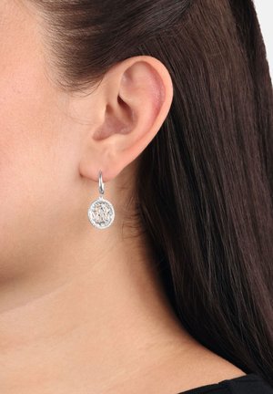 Pendiente de plata en forma de gota con un intrincado diseño de hojas, que lleva en la oreja izquierda de una persona con cabello castaño oscuro.