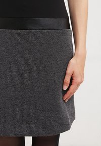 Šedá sukně s texturovanou tkaninou a černým koženým pasem. Má přiléhavý střih dosahující do poloviny stehna, s minimálními detaily.