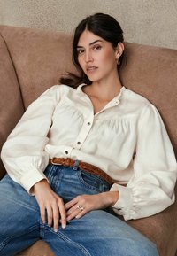 Crèmekleurige blouse met een geribbelde kraag en knoopsluiting, gecombineerd met hooggewaisted blauwe jeans en een geweven bruine leren riem.