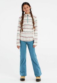 Multikleurige long sleeve zip-top met een geometrisch patroon in crème, gecombineerd met blauwe sneeuwbroeken en zwarte sneeuwlaarzen.
