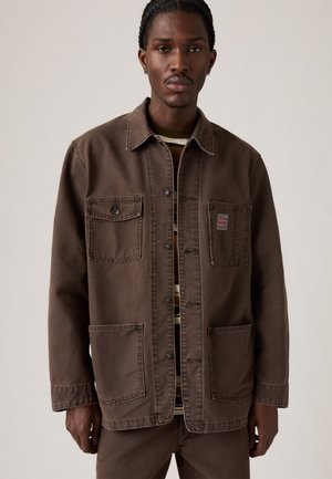 Homme avec des cheveux tressés portant une veste brune à boutons avec quatre poches par-dessus une chemise rayée, se tenant devant un fond uni.