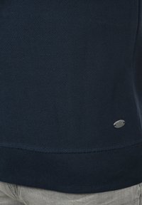 MARINFÄRGAD TEXTURERAD TRÖJA MED RIBBAD NEDRE KANT, FÖRSEDD MED EN METALLLOGO-KLISTERMÄRKE PÅ SIDAN. MATERIALITET SER MJUK OCH KNITTAD UT.