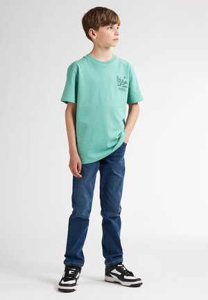 Lichtgroene katoenen t-shirt met korte mouwen, met een bergafbeelding op de borst. Gepaard met donkerblauwe spijkerbroek en zwarte sneakers.